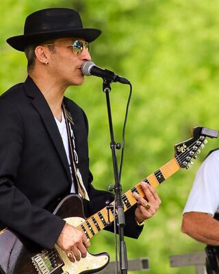 A musician in sunglasses and a black hat passionately plays guitar and sings into a microphone, with a bandmate visible nearby.