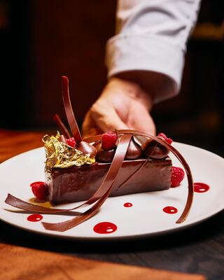 A decadent chocolate dessert garnished with gold foil, chocolate ribbons, and fresh raspberries, presented elegantly on a plate.