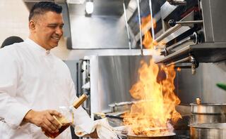 A chef joyfully adds a spirit to a sizzling pan, creating a vibrant flame in a bustling kitchen.