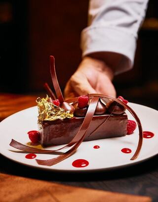 A luxurious chocolate dessert garnished with raspberries, chocolate curls, and gold leaf, elegantly presented on a white plate.