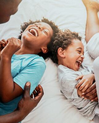 Two joyful children lie on a bed, laughing together, expressing pure happiness while an adult's hand gently touches one of them.