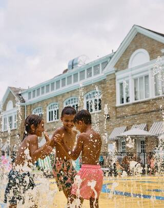 Three children play joyfully in a splash zone, surrounded by water fountains, with a lively background of a building and other kids.