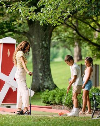 A woman watches as two children play miniature golf in a lush, green park. Bright sunlight filters through the trees, creating a cheerful scene.