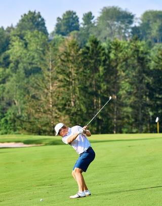 A golfer in a white shirt and shorts swings a club on a lush green course, surrounded by trees and under a blue sky.