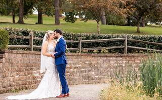 A couple in wedding attire shares a tender moment, framed by lush greenery and a rustic wooden fence in a serene outdoor setting.