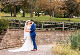 A couple in wedding attire shares a tender moment, framed by lush greenery and a rustic wooden fence in a serene outdoor setting.