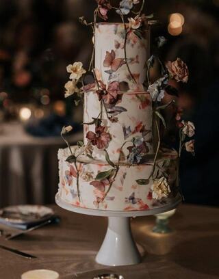 A three-tier cake adorned with delicate flowers and soft colors, elegantly displayed on a pedestal against a softly lit backdrop.
