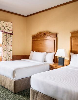 A cozy hotel room with two neatly made beds, wooden headboards, a bedside lamp, and a patterned curtain enhancing the warm ambiance.