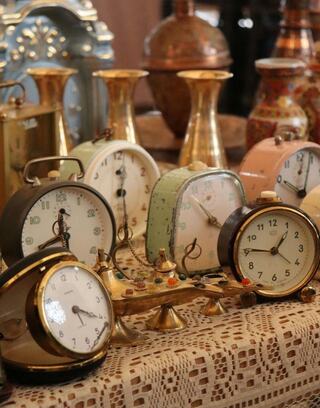 A collection of vintage alarm clocks in various colors and designs, displayed on a lace tablecloth amidst decorative vases.