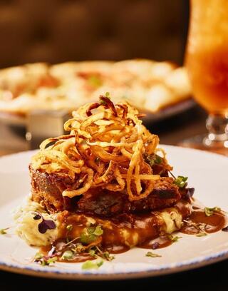 A hearty dish featuring tender meat topped with crispy onions, served on creamy mashed potatoes with a flavorful sauce and garnished with herbs.