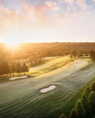 A serene golf course at sunrise, featuring rolling greens, sand traps, and surrounding trees bathed in warm light and soft shadows.