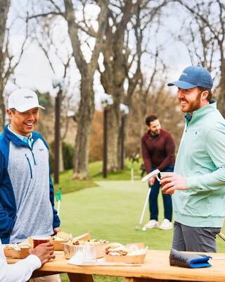 Friends enjoy food and laughter on a golf course, with one person practicing putts in the background during a relaxed gathering.