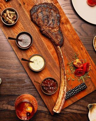 A large, juicy tomahawk steak rests on a wooden platter, accompanied by various sauces and garnishes for a flavorful feast.