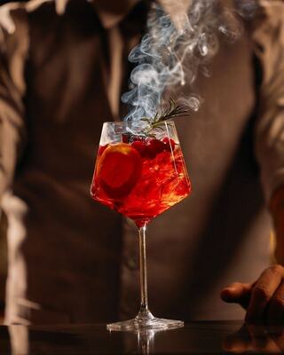 A vibrant cocktail in a stylish glass, garnished with herbs and citrus, emits swirling smoke, creating an enticing, mystic atmosphere.