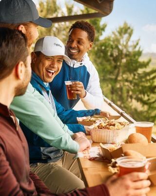 A group of friends enjoys food and drinks together on a patio, laughing and sharing good moments against a scenic backdrop of trees.