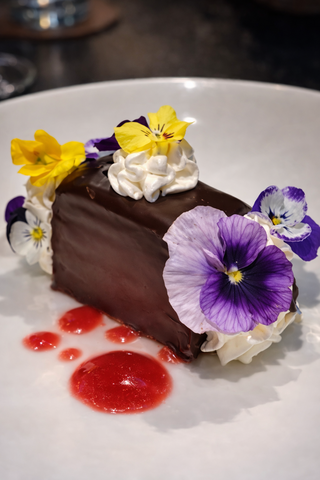 A luscious chocolate dessert adorned with vibrant edible flowers and swirls of whipped cream, complemented by a drizzle of strawberry sauce.