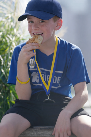 A young boy in a blue shirt and cap enjoys a snack while proudly wearing a medal, sitting against a sunny backdrop.