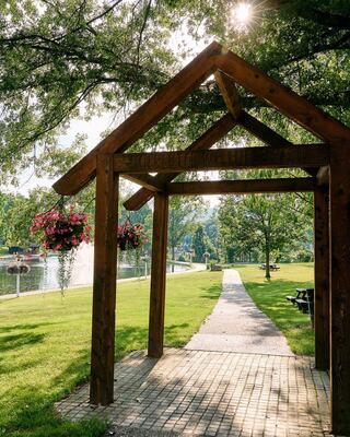 A wooden archway opens to a scenic park path lined with lush greenery and vibrant flower baskets, leading towards a tranquil body of water.