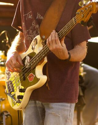 A musician plays a cream-colored bass guitar adorned with colorful stickers, wearing a casual maroon shirt and light jeans.