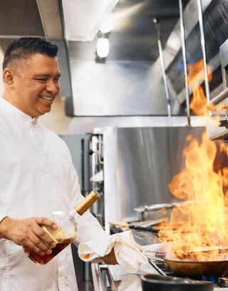 A smiling chef in a white coat stands in a busy kitchen, pouring a liquid into a flaming pan, surrounded by stainless steel appliances.