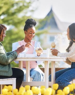 Three women enjoy ice cream together at an outdoor table, surrounded by greenery and blooming flowers, sharing laughter and smiles.