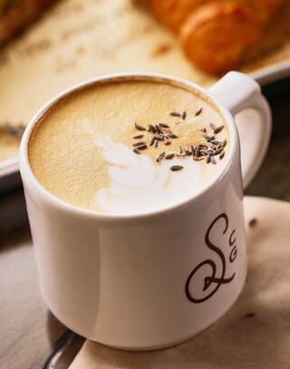 A creamy cup of coffee topped with lavender, featuring delicate latte art, beside a croissant on a rustic tray.