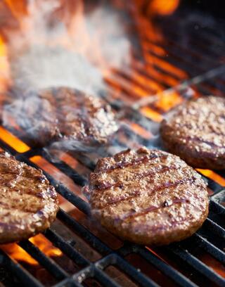 Sizzling burger patties on a grill, with flames licking and smoke rising, create a mouthwatering scene of outdoor cooking.