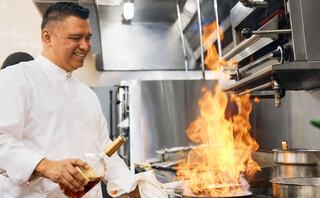 A chef joyfully prepares a dish in a bustling kitchen, with flames rising dramatically from the pan as he adds an ingredient.