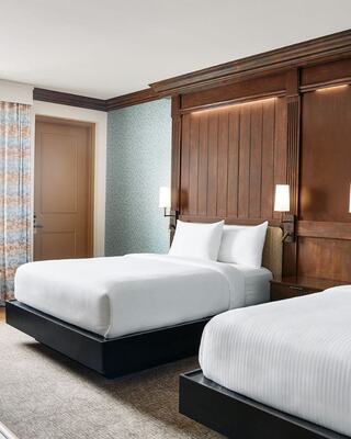 A cozy hotel room features two neatly made beds, warm wood accents, soft lighting, and a tasteful color palette, creating a welcoming atmosphere.