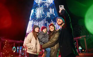 A joyful family takes a selfie in front of a brightly lit Christmas tree, surrounded by festive decorations and colorful lights.