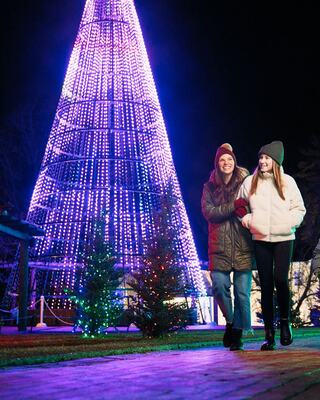 Two young women walk together, smiling, near a brightly lit Christmas tree adorned with colorful lights, creating a festive atmosphere.