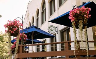 A charming café features blue umbrellas and vibrant pink flowers hanging from planters on a wooden deck, inviting outdoor dining.