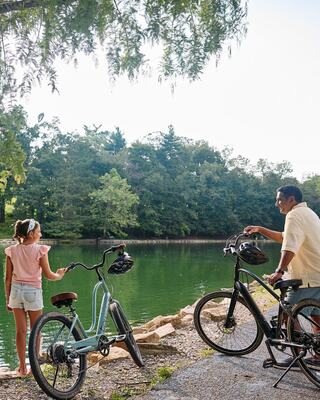 A girl and a man stand by a serene pond, chatting while their bicycles rest nearby, surrounded by lush greenery.