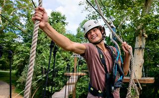 A smiling young man in a helmet navigates a rope course, showcasing excitement and confidence amid lush green trees.