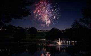 Vibrant fireworks burst over a tranquil lake, reflecting their colors in the water, while soft lights twinkle along the shore.