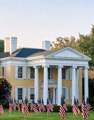 A grand yellow mansion features tall white columns, surrounded by numerous American flags on manicured grassy grounds under a clear sky.