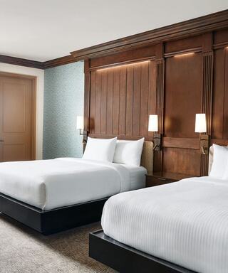 A stylish hotel room features two neatly made beds, a warm wooden accent wall, subtle lighting, and a calm, modern decor.