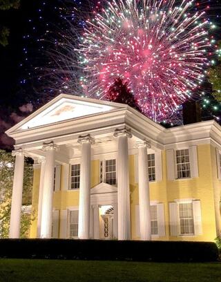 A grand yellow mansion with white columns illuminated at night, set against a vibrant display of colorful fireworks in the sky.