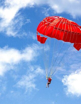 A person descends gracefully under a vibrant red parachute against a bright blue sky dotted with fluffy white clouds.