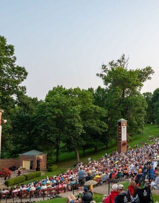 A large crowd gathers on a hillside for an outdoor event, surrounded by trees and an inviting stage area, creating a lively atmosphere.