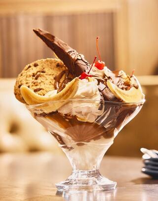 A decadent sundae features scoops of ice cream, chocolate sauce, bananas, cookies, and cherries, presented in an elegant glass dish.