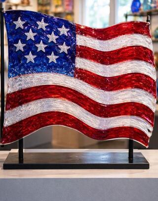 A glass sculpture of the American flag features vivid red and blue colors with swirling stripes and stars, set on a sleek black base.