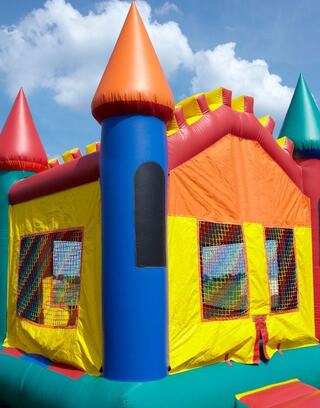 A colorful bounce house features bright orange and yellow sections with blue accents and pointed roofs, set against a clear blue sky.