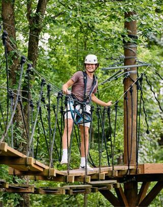 A young person in a helmet stands on a wooden rope bridge, smiling and harnessed, surrounded by lush green trees. Adventure awaits!