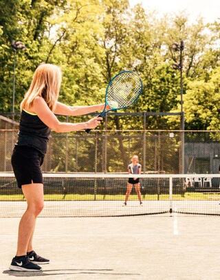 Two women play tennis on a sunny court, one preparing to serve while the other stands ready to receive. Lush green trees in the background.