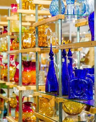 A display of colorful glassware in various shapes and sizes, showcasing vibrant hues of blue, orange, and red against a wooden shelf.