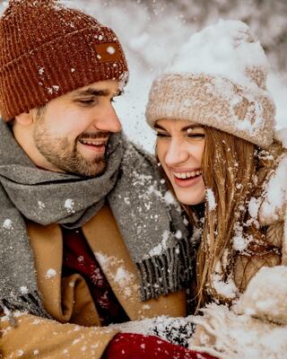 A smiling couple bundled in warm winter attire, surrounded by falling snow, joyfully sharing a cozy moment together.