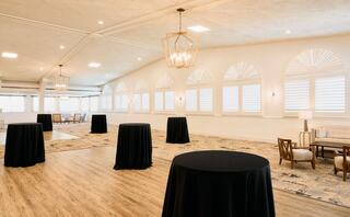 A spacious indoor venue features tables with black tablecloths, elegant chandeliers, and large windows allowing natural light.