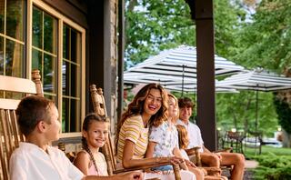 A cheerful woman and five children sit on rocking chairs, enjoying a sunny day, surrounded by greenery and striped umbrellas.