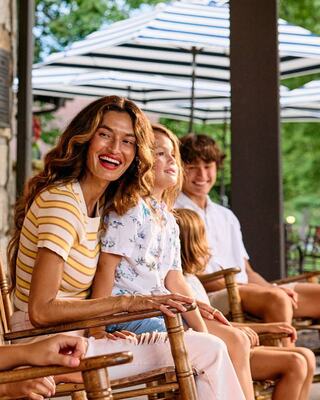 A woman with wavy hair and a bright smile sits among children, enjoying a sunny day on a porch with striped umbrellas in the background.